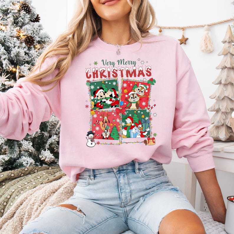 May include: A light pink sweatshirt with a festive Christmas design. The design features four squares with Disney characters and the text "Very Merry Christmas". The sweatshirt is a crew neck style.