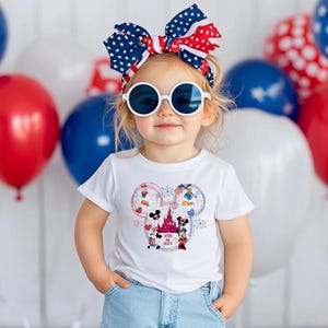 Peut inclure: Un enfant porte un t-shirt blanc avec un motif Mickey Mouse color&eacute; et le texte "4th of July". L'enfant porte &eacute;galement un bandeau et des lunettes de soleil patriotiques, avec des ballons rouges, blancs et bleus en arri&egrave;re-plan.