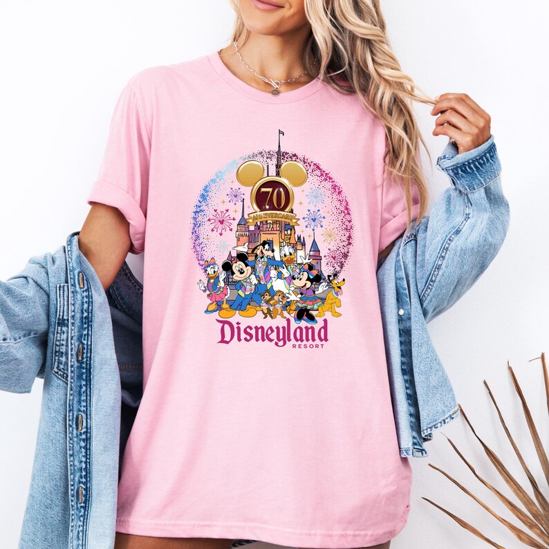 Op de afbeelding: Roze T-shirt met een afbeelding van Mickey Mouse en zijn vrienden die het 70-jarig jubileum van Disneyland vieren. De afbeelding bevat vuurwerk, het Disneyland-kasteel en de tekst "Disneyland Resort".
