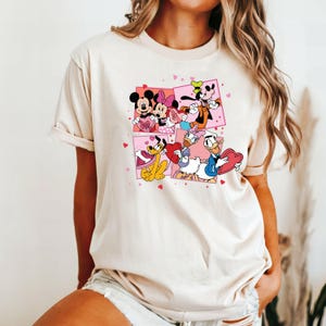 May include: A white t-shirt with a graphic print of Mickey Mouse, Minnie Mouse, Donald Duck, and Goofy holding hearts. The characters are surrounded by pink hearts.