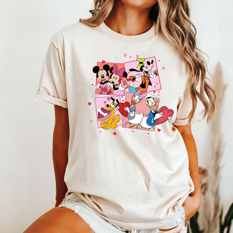 May include: A white t-shirt with a graphic print of Mickey Mouse, Minnie Mouse, Donald Duck, and Goofy holding hearts. The characters are surrounded by pink hearts.