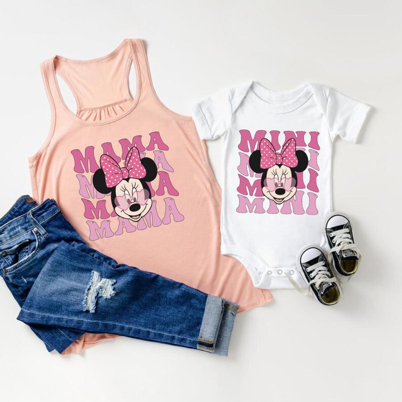 May include: A pink tank top with the text "MAMA" repeated in a pink and white pattern, featuring a pink and white Minnie Mouse graphic. A white bodysuit with the text "MINI" repeated in a pink and white pattern, featuring a pink and white Minnie Mouse graphic.