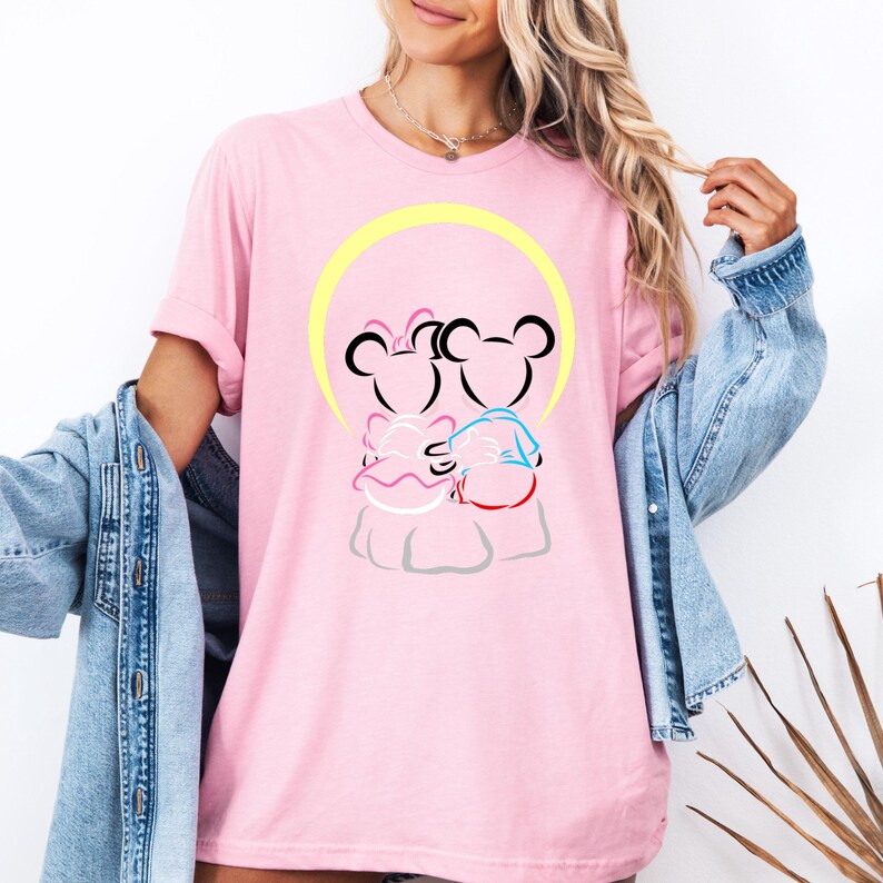 Puede incluir: Camiseta rosa claro con una ilustraci&oacute;n de dibujos animados de dos personajes abraz&aacute;ndose con orejas de rat&oacute;n, delineados en negro. El dise&ntilde;o incluye un halo amarillo y acentos de rosa, azul y rojo. La camiseta es de color rosa liso.