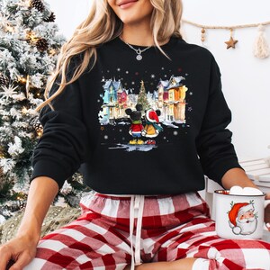 Op de afbeelding: Zwart sweatshirt met een aquarel illustratie van Mickey en Minnie Mouse in winterkleding, tegen een besneeuwd dorpsgezicht met een kerstboom. Het ontwerp bevat vallende sneeuwvlokken en kleurrijke huizen.