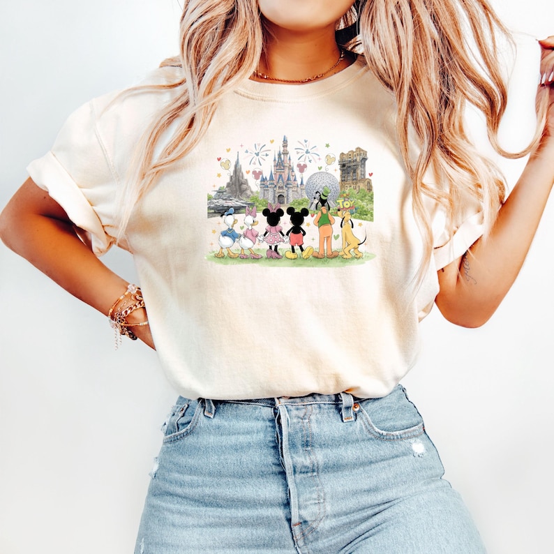 May include: Cream-colored t-shirt featuring a colorful illustration of Disney characters, including Mickey and Minnie Mouse, Donald and Daisy Duck, Goofy, and Pluto, in front of a castle and other park landmarks. The design includes fireworks and heart accents.