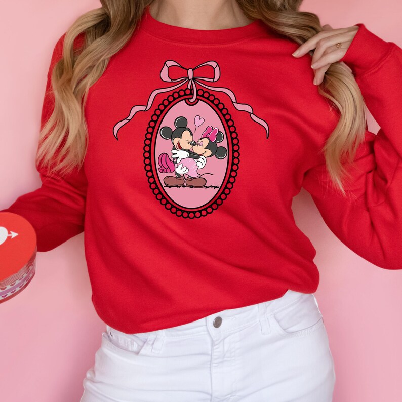 K&ouml;nnte beinhalten: Roter Pullover mit einer rosa Schleife und einem Motiv von Mickey Mouse und Minnie Mouse, die sich in einem rosa ovalen Rahmen k&uuml;ssen.