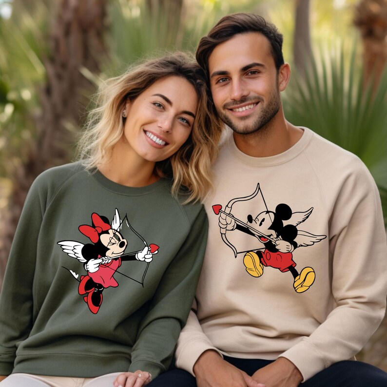 May include: A couple wearing matching green and beige sweatshirts with a cartoon image of Mickey Mouse and Minnie Mouse dressed as cupids. The sweatshirts feature a red heart and arrow design.