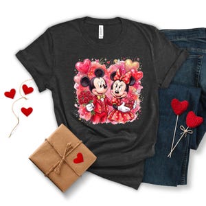 May include: Dark gray t-shirt featuring a watercolor illustration of Mickey and Minnie Mouse in Valentine's Day attire. Mickey holds roses, and Minnie holds a heart-shaped gift. The design includes heart-shaped balloons and a pink and red background.