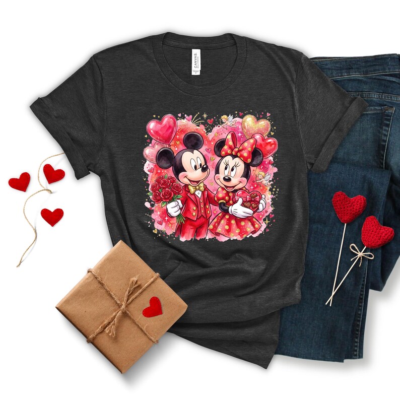 May include: Dark gray t-shirt featuring a watercolor illustration of Mickey and Minnie Mouse in Valentine's Day attire. Mickey holds roses, and Minnie holds a heart-shaped gift. The design includes heart-shaped balloons and a pink and red background.