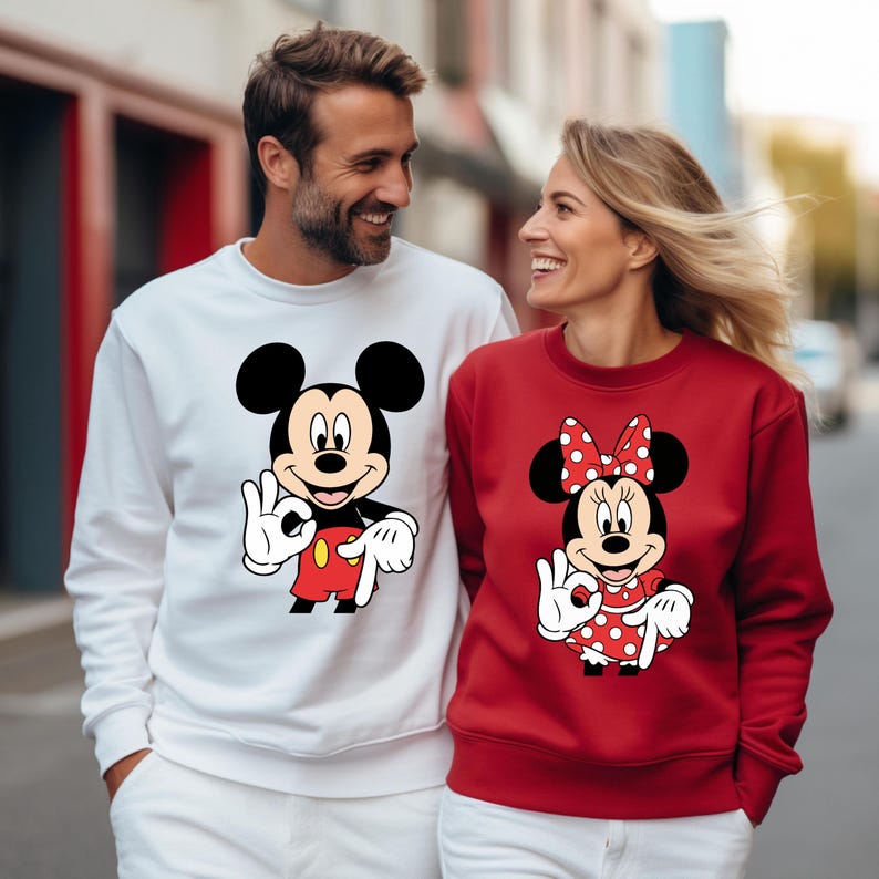 May include: Matching white and red sweatshirts featuring Mickey and Minnie Mouse. Mickey is on a white sweatshirt, wearing red shorts and giving an 'okay' hand gesture. Minnie is on a red sweatshirt, wearing a polka dot dress and bow.