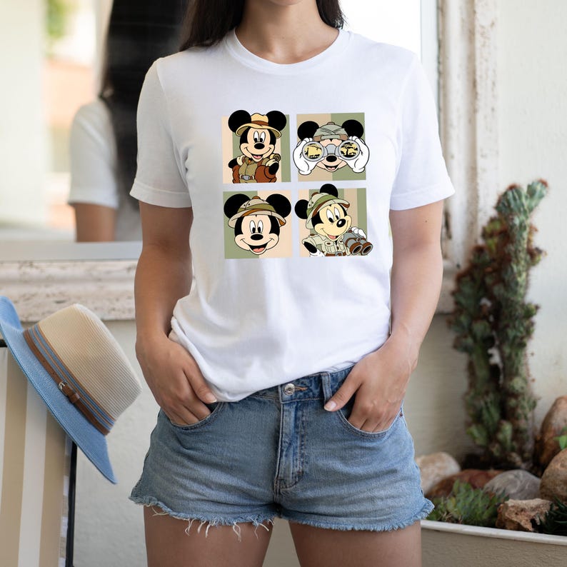 K&ouml;nnte beinhalten: Wei&szlig;es T-Shirt mit vier Panels, die Micky Maus in Safari-Kleidung zeigen. Jedes Panel zeigt eine andere Pose von Micky, der einen Safarihut und -outfit tr&auml;gt, mit Fernglas und Kamera. Der Hintergrund ist eine Mischung aus Gr&uuml;n, Beige und Oliv.