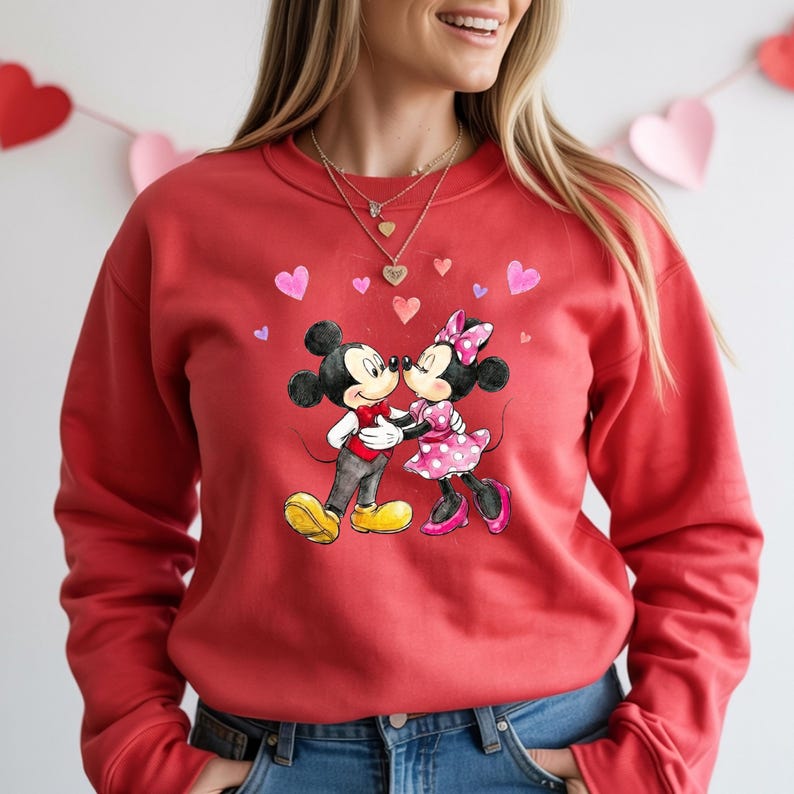 May include: Red sweatshirt with a watercolour illustration of Mickey and Minnie Mouse kissing, surrounded by pink hearts. Mickey wears a red bow tie and yellow shoes, Minnie a pink polka-dot dress and bow. A romantic design.