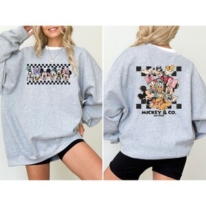 May include: A heather gray sweatshirt featuring a colorful graphic of Disney characters, including Mickey Mouse, Minnie Mouse, and Donald Duck. The front graphic is a row of characters above a black and white checkered pattern. The back graphic features a group of characters with the text "Mickey & Co. EST 1928".
