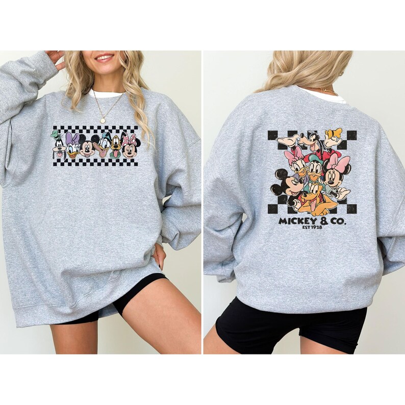 May include: A heather gray sweatshirt featuring a colorful graphic of Disney characters, including Mickey Mouse, Minnie Mouse, and Donald Duck. The front graphic is a row of characters above a black and white checkered pattern. The back graphic features a group of characters with the text "Mickey & Co. EST 1928".