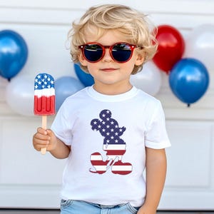 May include: A child wearing red sunglasses and a white t-shirt featuring a Mickey Mouse silhouette in the American flag design. The child holds a red, white, and blue ice lolly. Red, white, and blue balloons are in the background.