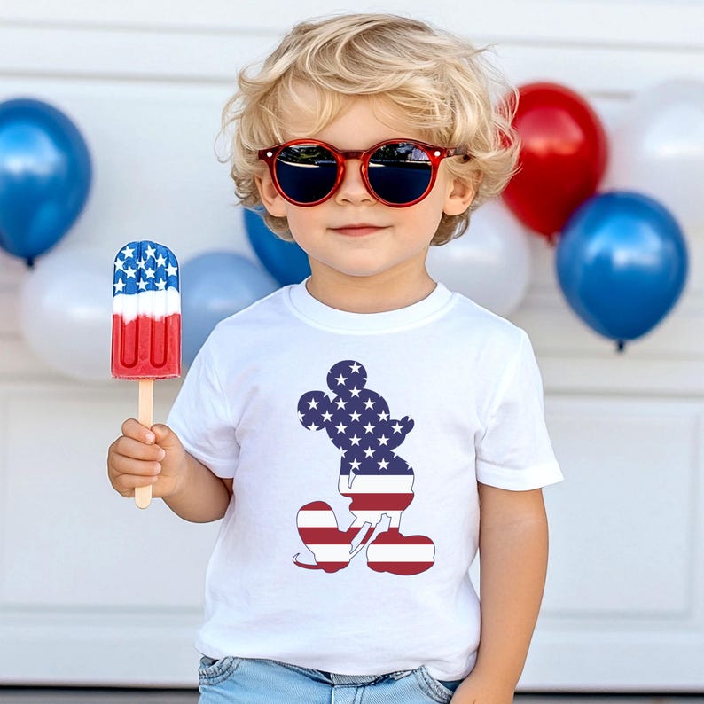 May include: A child wearing red sunglasses and a white t-shirt featuring a Mickey Mouse silhouette in the American flag design. The child holds a red, white, and blue ice lolly. Red, white, and blue balloons are in the background.
