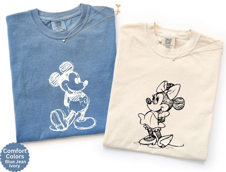 May include: Two Comfort Colors t-shirts, one blue jean and one ivory, each with a sketched cartoon character. The blue shirt features Mickey Mouse, and the ivory shirt features Minnie Mouse. Both shirts have a small gold necklace.