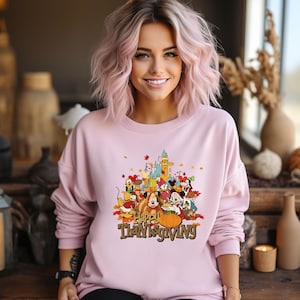 May include: A light pink sweatshirt featuring a colorful graphic of cartoon characters and a castle, with the text "Happy Thanksgiving". The design includes a pumpkin and autumn leaves. The sweatshirt is worn by a person with pink hair.