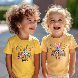 May include: Two children wearing yellow t-shirts with a colorful Disney Trip 2025 graphic. The shirts feature Mickey Mouse ears with fireworks and a group of Disney characters. One shirt has a pink bow on the ears.