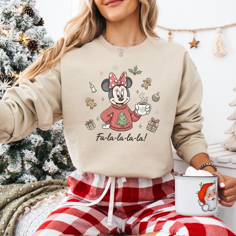 May include: Beige sweatshirt featuring a Minnie Mouse graphic in a red sweater with a Christmas tree design, holding a mug. The design includes gingerbread men, gifts, and the text "Fa-la-la-la-la!".
