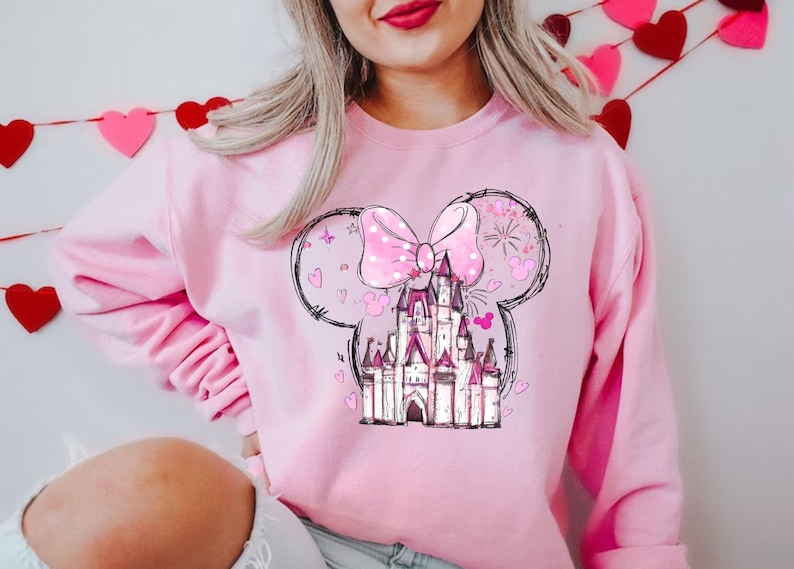 May include: Pink sweatshirt with a graphic of a pink castle with a pink bow and polka dots, surrounded by hearts and stars. The graphic is in the shape of Mickey Mouse ears.