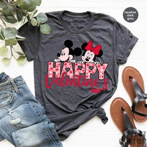May include: Heather dark grey t-shirt featuring a graphic of Mickey Mouse and Minnie Mouse peeking over the text "HAPPY VALENTINE'S DAY" with pink hearts.
