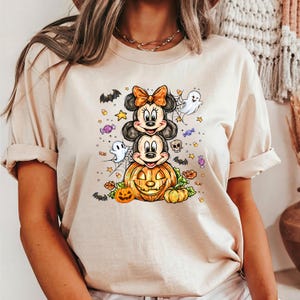 May include: Beige t-shirt with a Halloween design. The graphic shows Mickey and Minnie Mouse characters, a carved pumpkin, bats, ghosts, and sweets. The shirt is made of a soft material. A fun, seasonal top.