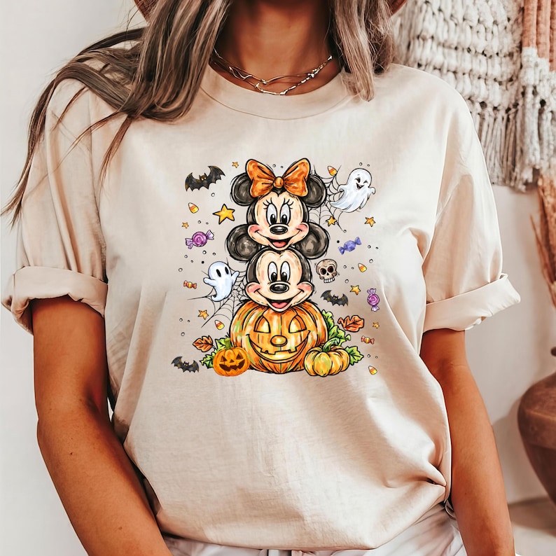 May include: Beige t-shirt with a Halloween design. The graphic shows Mickey and Minnie Mouse characters, a carved pumpkin, bats, ghosts, and sweets. The shirt is made of a soft material. A fun, seasonal top.