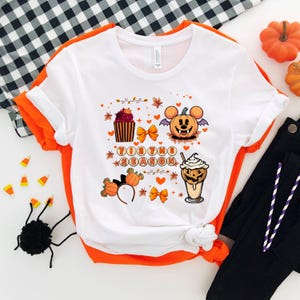 Puede incluir: Camiseta blanca con ribete naranja con un gr&aacute;fico tem&aacute;tico de Halloween. El dise&ntilde;o incluye una calabaza con orejas de rat&oacute;n, una magdalena, una bebida y el texto "Tis the Season". La camiseta se combina con pantalones negros.