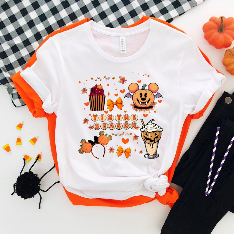 Puede incluir: Camiseta blanca con ribete naranja con un gr&aacute;fico tem&aacute;tico de Halloween. El dise&ntilde;o incluye una calabaza con orejas de rat&oacute;n, una magdalena, una bebida y el texto "Tis the Season". La camiseta se combina con pantalones negros.