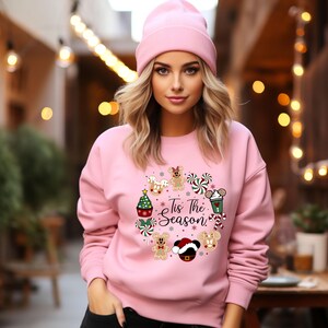 Peut inclure: Un ensemble rose compos&eacute; d'un sweat-shirt et d'un bonnet. Le sweat-shirt pr&eacute;sente un motif de couronne de No&euml;l avec des bonhommes en pain d'&eacute;pices, des cannes en sucre et un bonnet de P&egrave;re No&euml;l. Le texte "Tis The Season" est au centre.