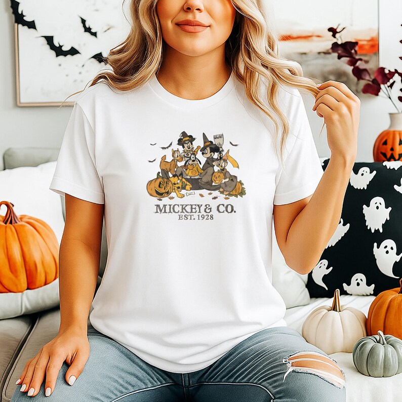 Pode incluir: Camiseta branca com um gr&aacute;fico de Halloween de personagens de desenhos animados, ab&oacute;boras e morcegos. O texto "Mickey & Co. Est. 1928" est&aacute; impresso abaixo. A camisa &eacute; de gola redonda.