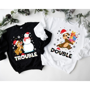 Puede incluir: Dos sudaderas con tem&aacute;tica navide&ntilde;a, una negra con un mu&ntilde;eco de nieve y una ardilla con el texto "Trouble" y una blanca con una ardilla llevando regalos y el texto "Double".