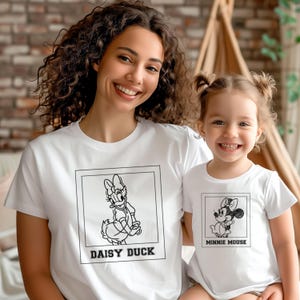 May include: Two white t-shirts featuring Disney characters. One shirt displays Daisy Duck, the other Minnie Mouse. Both shirts have a black and white graphic design within a rectangular frame. The shirts are worn by a woman and a child.