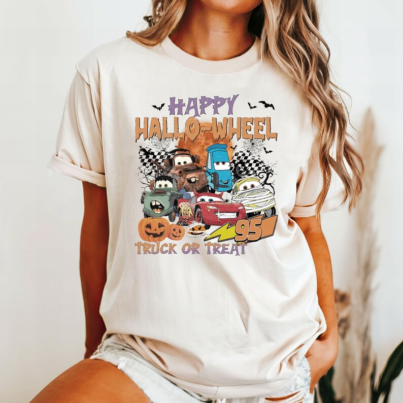 May include: A beige t-shirt with a graphic of cartoon cars and pumpkins. The text "Happy Hallo-Wheel Truck or Treat" is printed on the shirt.