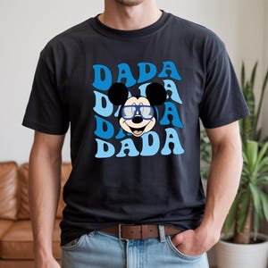 K&ouml;nnte beinhalten: Schwarzes T-Shirt mit einem blauen Retro-Design, das dreimal das Wort "DADA" zeigt. In der Mitte befindet sich eine Cartoon-Mickey-Mouse mit Brille. Dieses l&auml;ssige T-Shirt ist ein Alltagsartikel.
