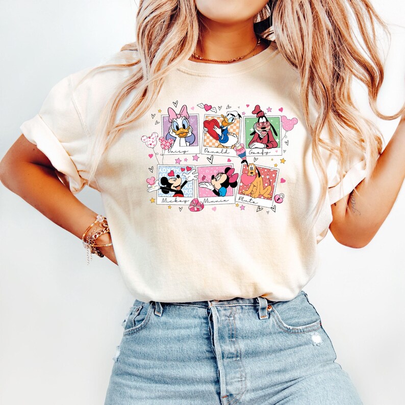 K&ouml;nnte beinhalten: Ein wei&szlig;es T-Shirt mit einem Grafikdruck von Mickey Mouse, Minnie Mouse, Donald Duck, Daisy Duck, Goofy und Pluto. Die Figuren sind in einer Collage von Polaroid-artigen Rahmen mit Herzen und Sternen angeordnet. Der Text "Mickey", "Minnie", "Donald", "Daisy", "Goofy" und "Pluto" steht unter jeder Figur.