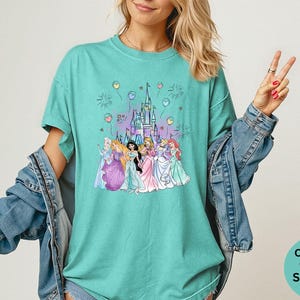 K&ouml;nnte beinhalten: T&uuml;rkisgr&uuml;nes T-Shirt mit einer Grafik eines Schlosses und Prinzessinnen, Ballons und Feuerwerk. Das T-Shirt ist mit "Comfort Colors Seafoam" beschriftet.