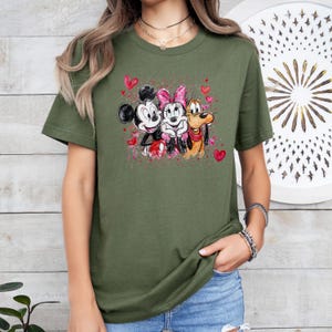 May include: Olive green t-shirt featuring a watercolor design of Mickey Mouse, Minnie Mouse, and Pluto with hearts. Mickey Mouse is wearing a black hat and red shorts. Minnie Mouse has a pink bow. Pluto has a red collar.