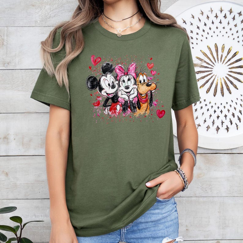 May include: Olive green t-shirt featuring a watercolor design of Mickey Mouse, Minnie Mouse, and Pluto with hearts. Mickey Mouse is wearing a black hat and red shorts. Minnie Mouse has a pink bow. Pluto has a red collar.