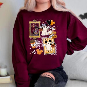 Puede incluir: Sudadera burdeos con gr&aacute;ficos tem&aacute;ticos de Halloween. El dise&ntilde;o incluye im&aacute;genes enmarcadas de un castillo embrujado, Mickey y Minnie Mouse disfrazados de fantasmas, una taza de caf&eacute;, calabazas y telara&ntilde;as. Tambi&eacute;n son visibles las palabras "Minnie Magic".