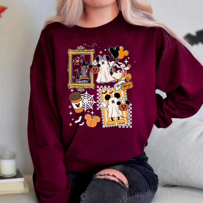 Puede incluir: Sudadera burdeos con gr&aacute;ficos tem&aacute;ticos de Halloween. El dise&ntilde;o incluye im&aacute;genes enmarcadas de un castillo embrujado, Mickey y Minnie Mouse disfrazados de fantasmas, una taza de caf&eacute;, calabazas y telara&ntilde;as. Tambi&eacute;n son visibles las palabras "Minnie Magic".
