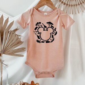 May include: A peach-coloured baby onesie with short sleeves features a black silhouette of a Mickey Mouse head. The silhouette is made up of various animals and plants, including a giraffe, a zebra, and trees.