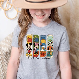 May include: A gray t-shirt featuring Mickey Mouse, Minnie Mouse, Donald Duck, and Goofy in colorful rectangular designs. The phrase "back to school" is printed in black script below the characters. The shirt is worn by a person.