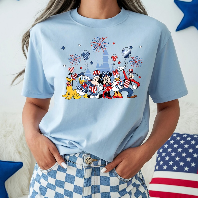 K&ouml;nnte beinhalten: Hellblaues T-Shirt mit einem patriotischen Design mit Zeichentrickfiguren, Feuerwerk und Luftballons. Die Grafik zeigt Micky Maus, Minnie Maus, Donald Duck und Goofy in rot, wei&szlig; und blau. Das Shirt hat einen Rundhalsausschnitt und kurze &Auml;rmel.