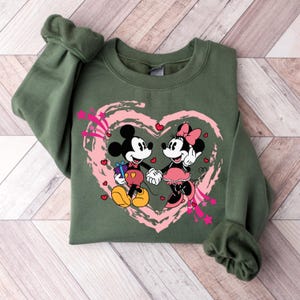 May include: An olive green sweatshirt featuring a graphic of Mickey and Minnie Mouse holding hands inside a pink heart. Mickey Mouse is holding a gift. The design includes pink stars and hearts.