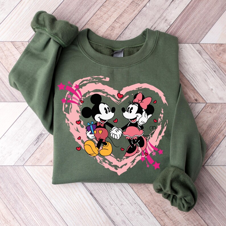 May include: An olive green sweatshirt featuring a graphic of Mickey and Minnie Mouse holding hands inside a pink heart. Mickey Mouse is holding a gift. The design includes pink stars and hearts.