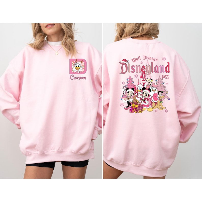 May include: A light pink sweatshirt featuring a custom design with a cartoon duck and the word "Custom". The back of the sweatshirt displays the text "Walt Disney's Disneyland 1955" with cartoon characters and holiday accents.