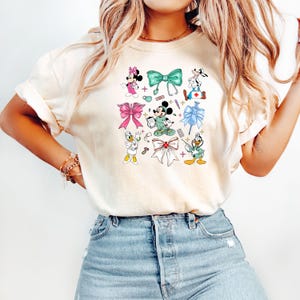 Pu&ograve; includere: T-shirt color crema con una grafica colorata di personaggi Disney in abiti medici e fiocchi. Minnie Mouse, Topolino, Pippo e Paperino sono raffigurati con strumenti medici e fiocchi in vari colori, tra cui rosa, verde e blu.