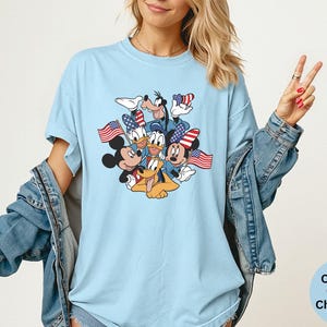 May include: Light blue t-shirt featuring a graphic of Mickey Mouse, Minnie Mouse, Donald Duck, Daisy Duck, Goofy, and Pluto, each holding an American flag. The shirt is a comfort colors chambray.